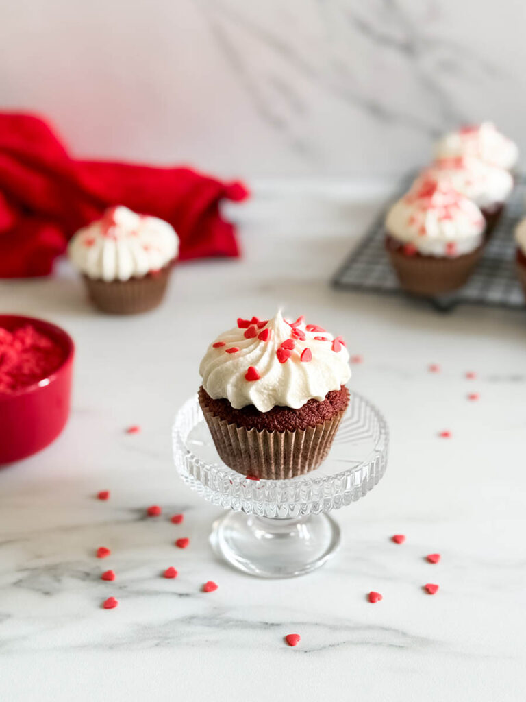Baileys Red Velvet Cupcakes, The Perfect Valentine's Dessert 8 A red velvet cupcake with heart-shaped sprinkles, sitting on a glass stand, styled with scattered red sprinkles, a red napkin, and additional cupcakes in the scene.