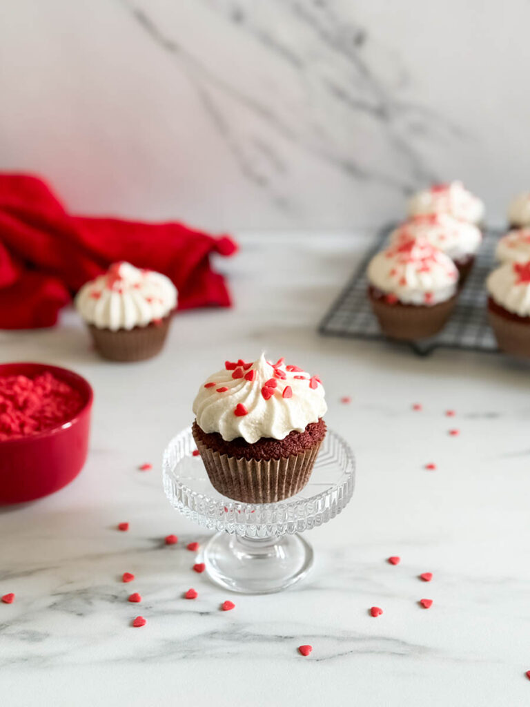 Baileys Red Velvet Cupcakes, The Perfect Valentine's Dessert 10 A red velvet cupcake with heart-shaped sprinkles, sitting on a glass stand, styled with scattered red sprinkles, a red napkin, and additional cupcakes in the scene.