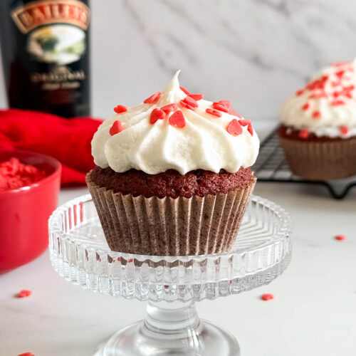close up red velvet cupcake on a cake stand, more cupcakes in the background with a red towel and a wire rack, more sugar hearts around, on a marble background, bottle of baileys