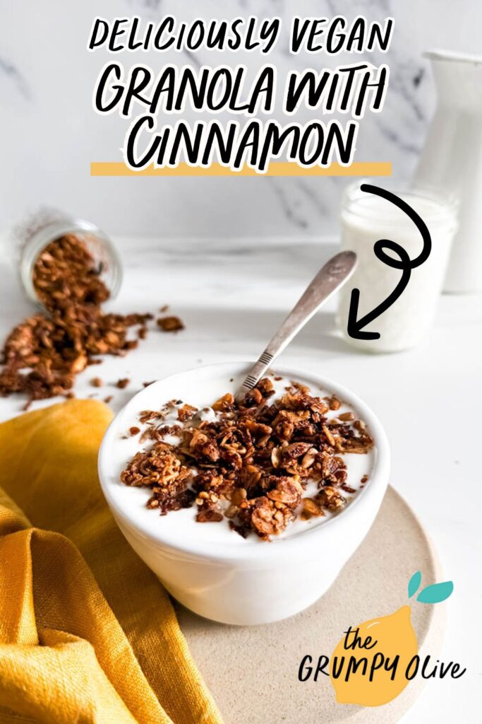 A bowl of vegan granola with cinnamon atop creamy yogurt, garnished with crunchy clusters, placed on a beige plate. A yellow cloth and a spilled jar of granola add a rustic touch, with milk in the background.