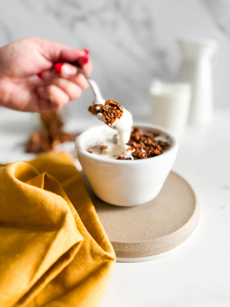 A close-up of a hand holding a spoonful of granola with cinnamon and yogurt, with a vibrant yellow cloth and granola jar in the background.