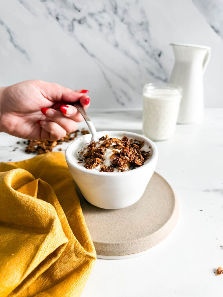 A hand lifting a spoon from a bowl of granola with cinnamon and yogurt. The scene includes a yellow cloth and a jar of granola with milk in the background on a marble surfa