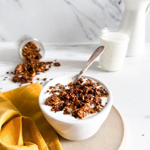Granola with cinnamon in a bowl of creamy yogurt, placed on a simple beige plate. A vibrant yellow cloth and spilled granola enhance the cozy breakfast vibe, with a milk jar in the background.