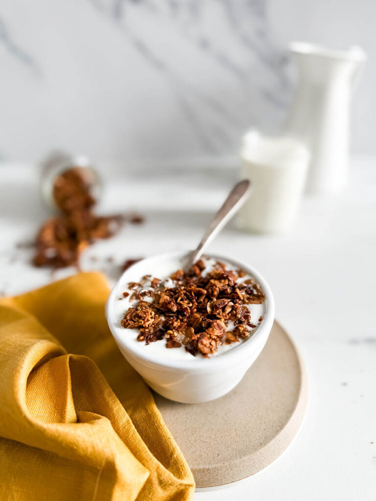 Granola with cinnamon in a bowl of creamy yogurt, placed on a simple beige plate. A vibrant yellow cloth and spilled granola enhance the cozy breakfast vibe, with a milk jar in the background.
