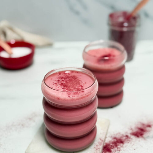 Red Latte Recipe, One Perfect For Valentine's Or Any Other Day Really 5 A creamy red latte topped with a dusting of red powder, served in a stackable glass on a white marble coaster. The background features a blurred jar of powder and a red bowl, adding to the cozy vibe.