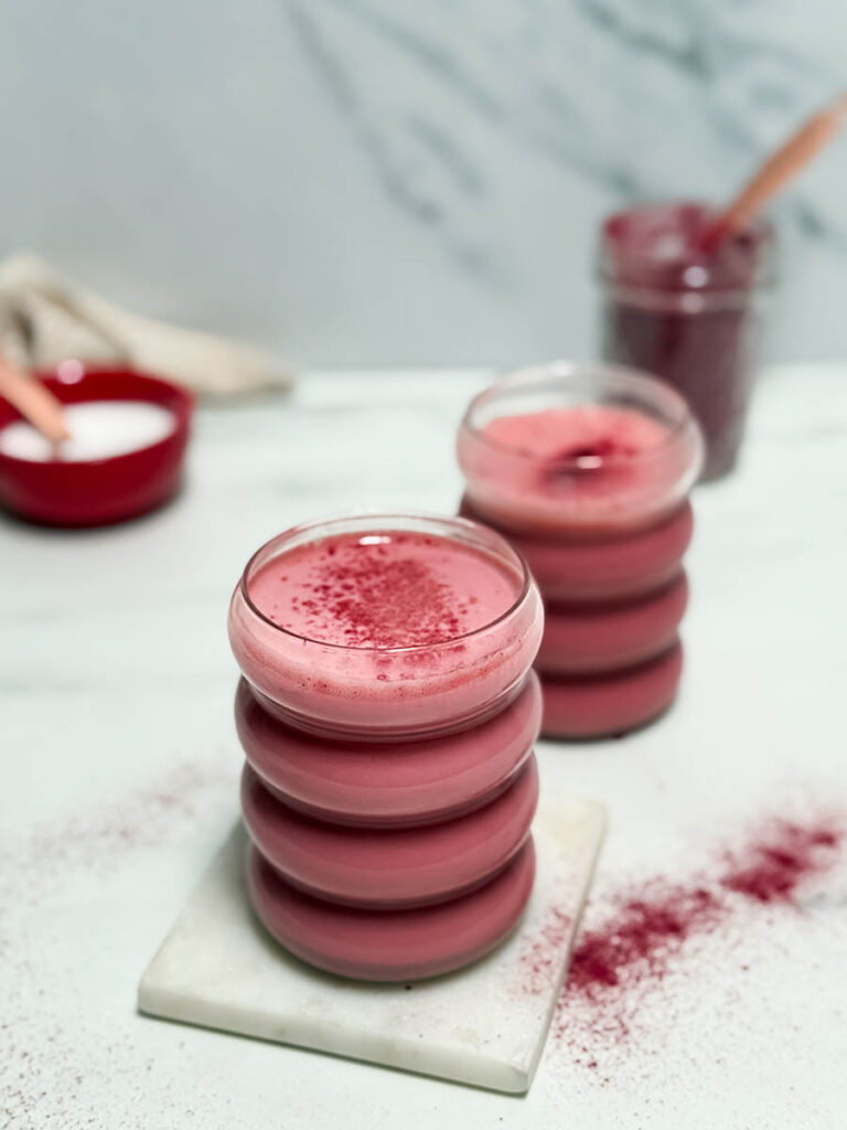 Red Latte Recipe, One Perfect For Valentine's Or Any Other Day Really 1 A creamy red latte topped with a dusting of red powder, served in a stackable glass on a white marble coaster. The background features a blurred jar of powder and a red bowl, adding to the cozy vibe.