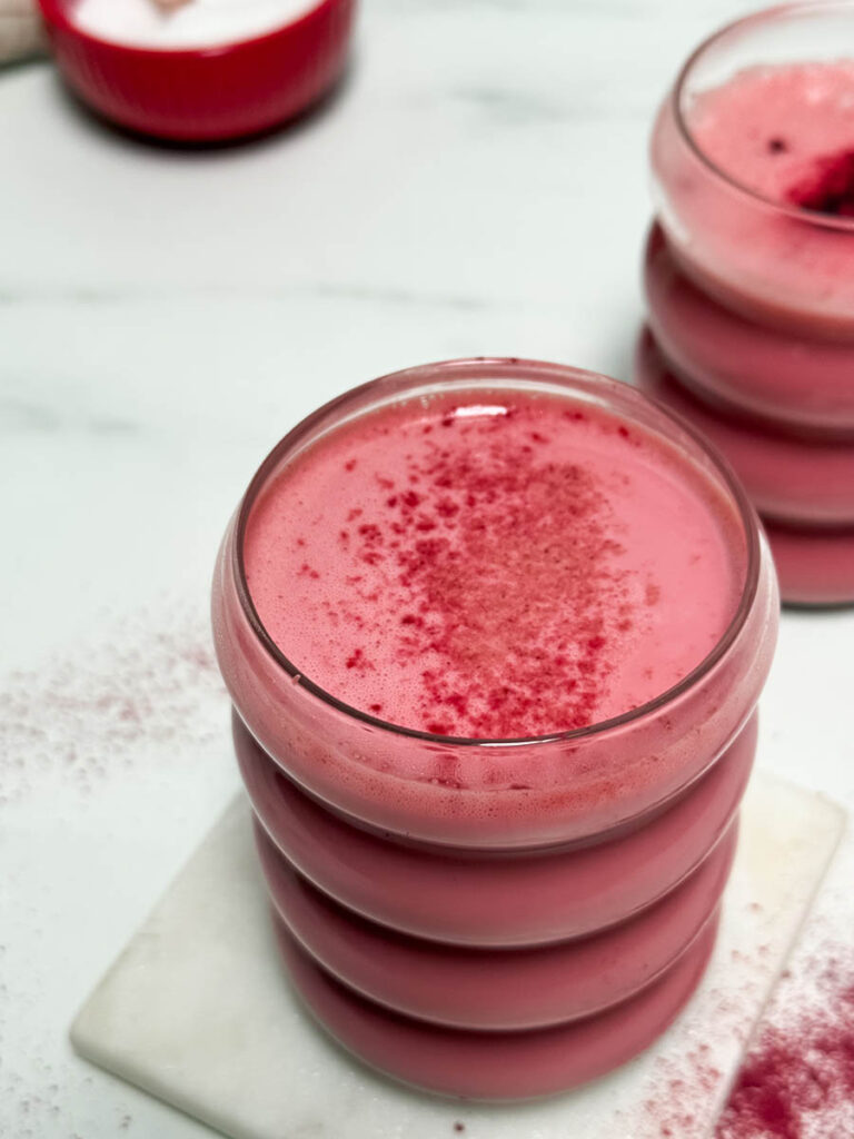 Red Latte Recipe, One Perfect For Valentine's Or Any Other Day Really 4 Close-up of a red latte in a ribbed glass, showcasing the frothy texture and dusted red powder topping. A red bowl and fabric napkin are softly visible in the background.