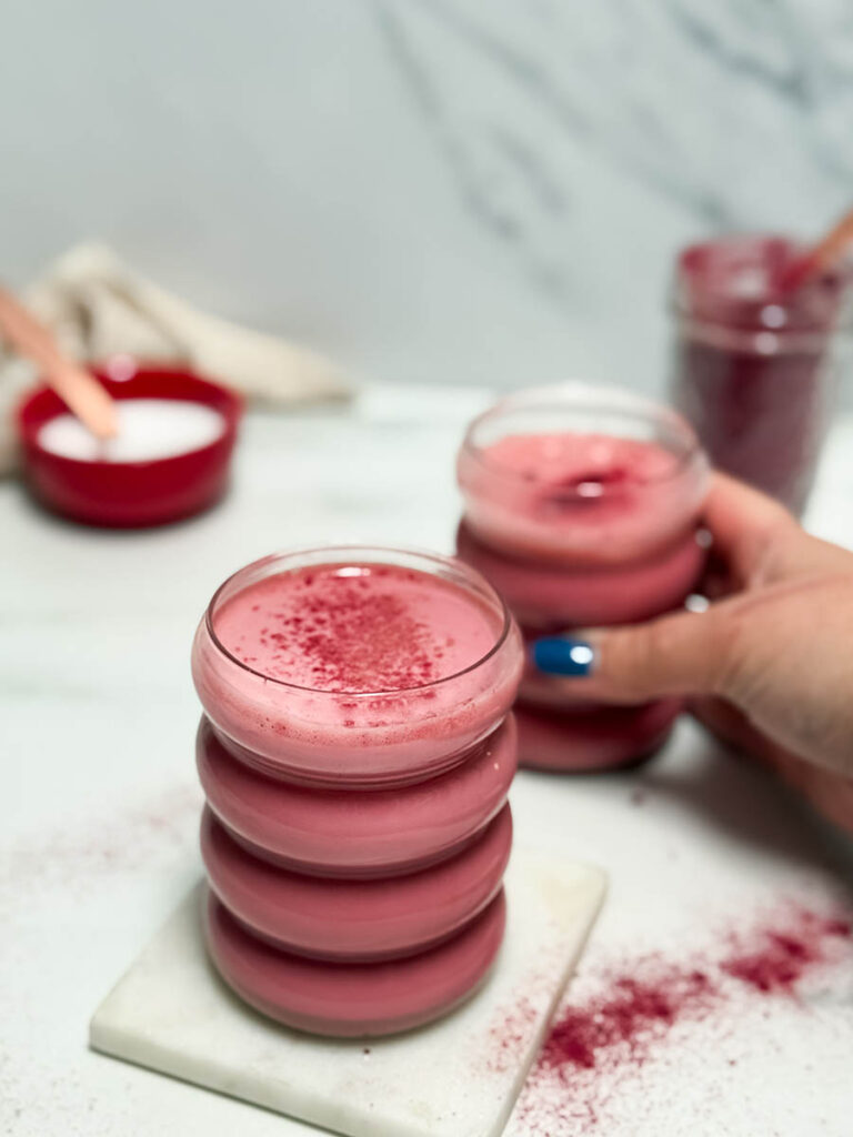 Red Latte Recipe, One Perfect For Valentine's Or Any Other Day Really 3 A person holding a glass of red latte, emphasizing its stacked design and creamy texture. The blurred backdrop includes another latte and a jar with a spoon, highlighting a relaxed, homemade atmosphere.