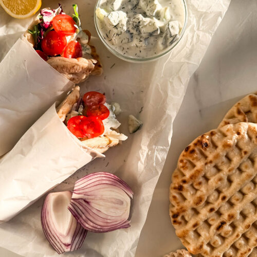 Top-down view of chicken pitas on parchment paper, filled with grilled chicken, fresh cherry tomatoes, mixed greens, and drizzled with creamy tzatziki sauce. Surrounding the wraps are a bowl of tzatziki, red onion slices, pita bread, and fresh lemon halves.