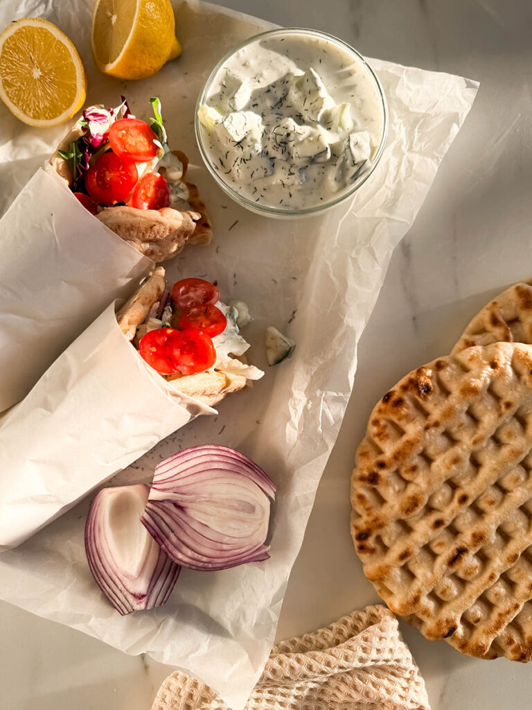 Simple Chicken Pitas, The Greek Way: Delicious And Quick 1 Top-down view of chicken pitas on parchment paper, filled with grilled chicken, fresh cherry tomatoes, mixed greens, and drizzled with creamy tzatziki sauce. Surrounding the wraps are a bowl of tzatziki, red onion slices, pita bread, and fresh lemon halves.