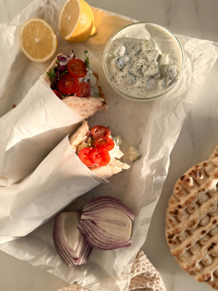 Simple Chicken Pitas, The Greek Way: Delicious And Quick 4 A fresh Mediterranean spread featuring chicken pitas, with bright tomatoes and leafy greens peeking out from the soft pita wrap. A lemon cut in half and a bowl of tzatziki sauce with dill complement the meal.