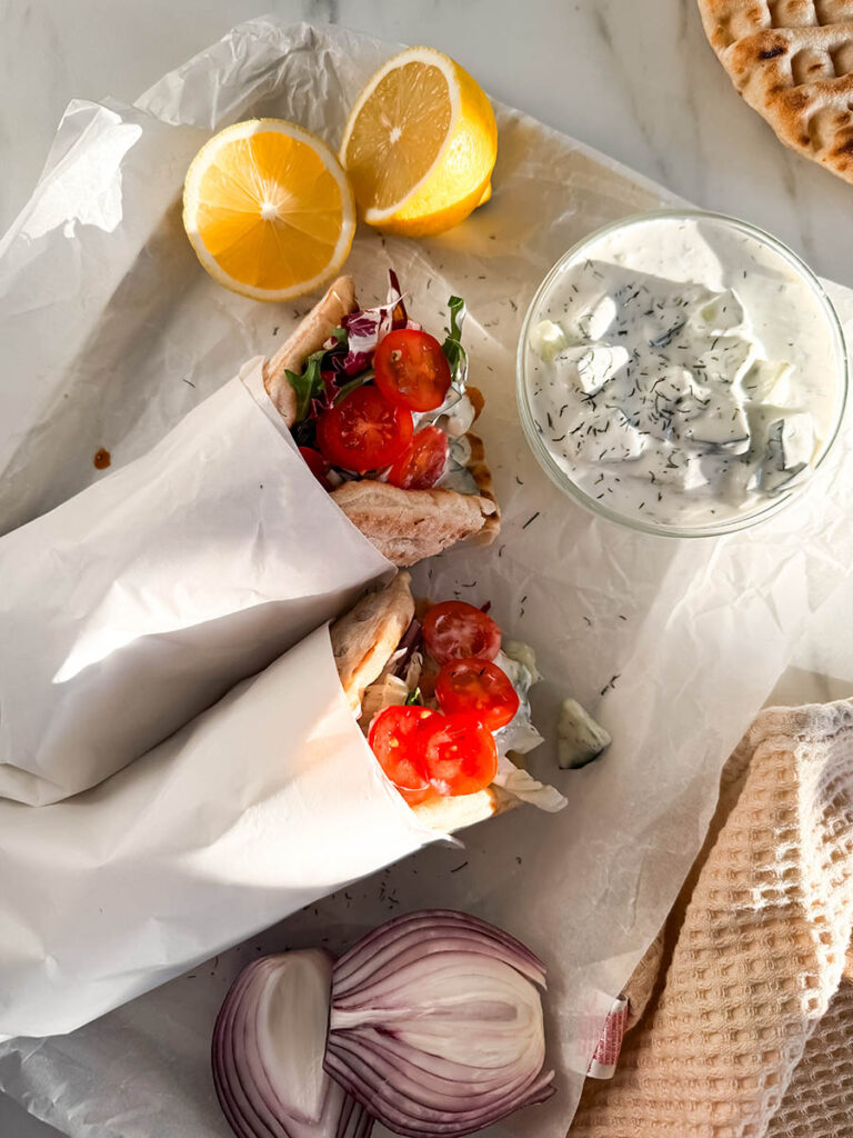 Simple Chicken Pitas, The Greek Way: Delicious And Quick 7 Chicken pitas served with classic Mediterranean sides, including creamy tzatziki, grilled pita bread, fresh red onion, and lemon wedges. The paper-wrapped pitas are neatly arranged on a parchment-lined surface.