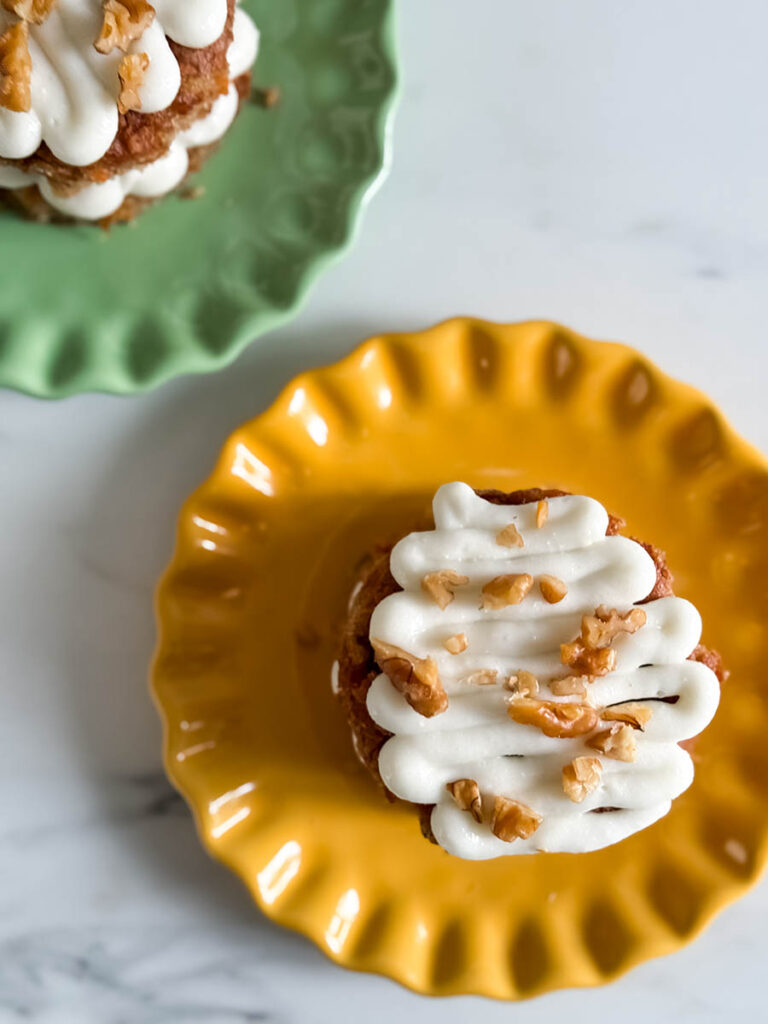 Top-down view of a mini carrot cake on a yellow scalloped plate, drizzled with piped cream cheese frosting and topped with chopped walnuts. A second cake is partially visible on a green plate in the background.