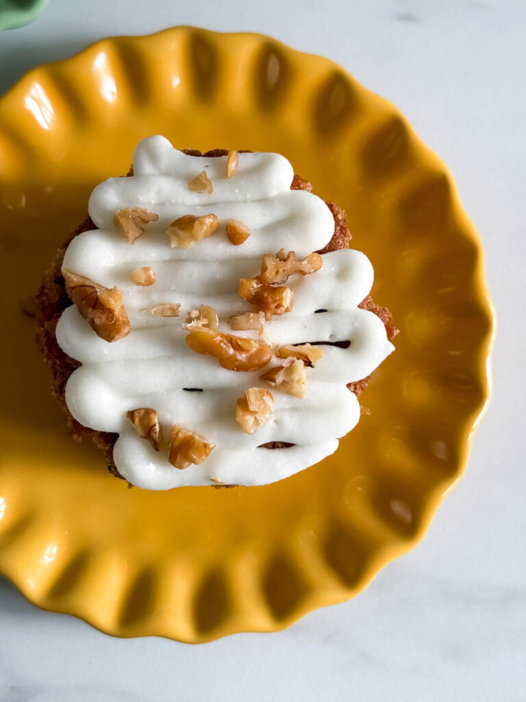 Close-up of a mini carrot cake with thick cream cheese frosting in a zig-zag pattern, sprinkled with walnuts, sitting on a bright yellow plate.