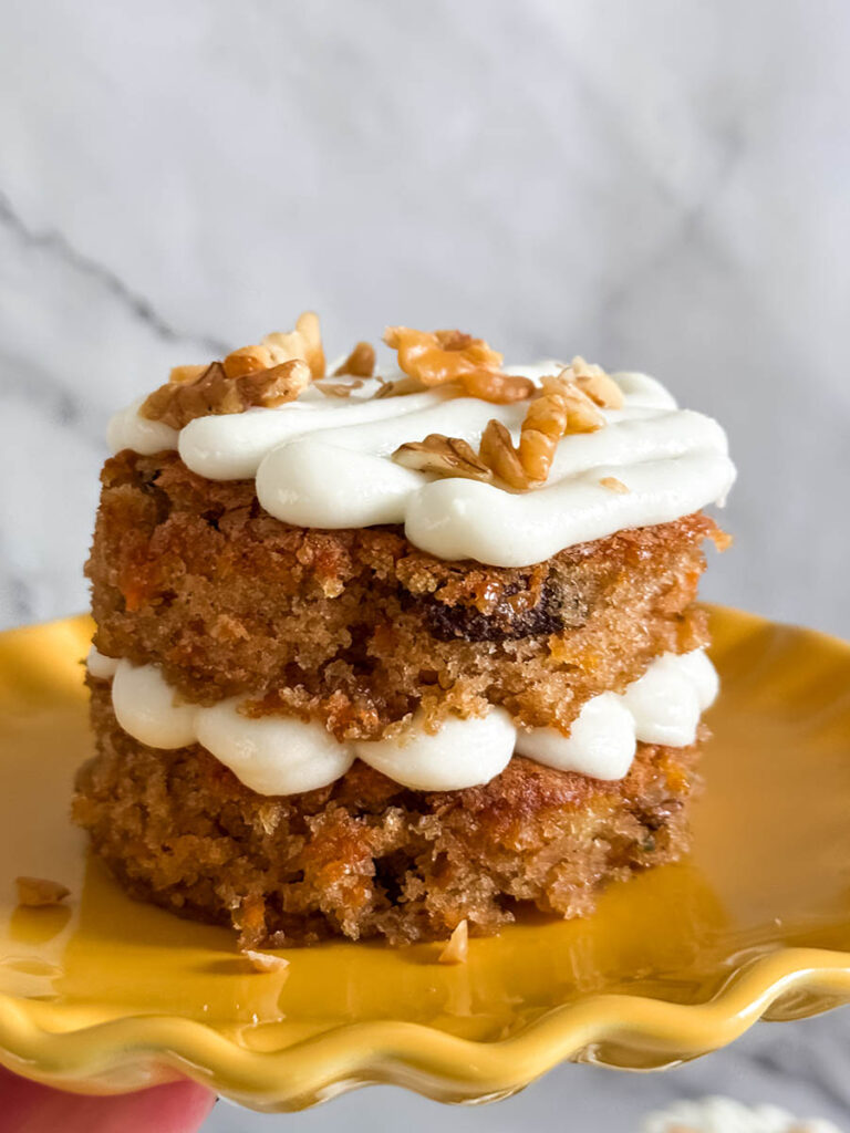 Side view of a two-layer mini carrot cake with visible shredded carrots and pecans, stacked with piped cream cheese frosting and garnished with walnuts.