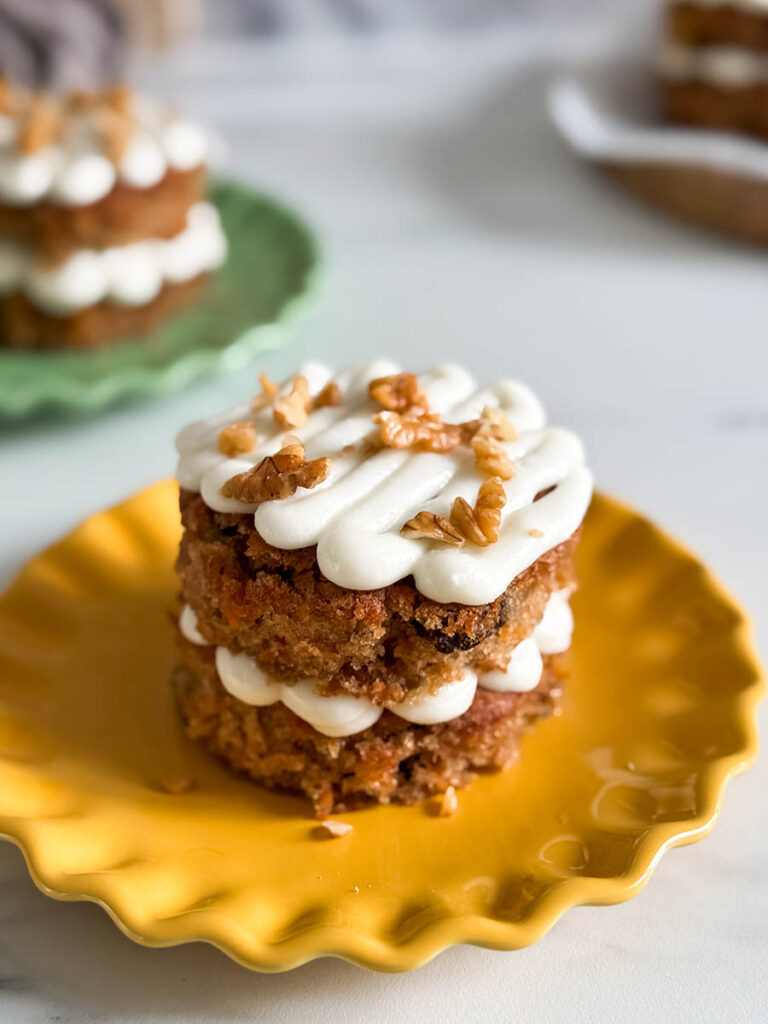 Another angle of a stacked carrot cake with creamy frosting and walnuts, placed on a yellow plate, with additional cakes blurred in the background.
