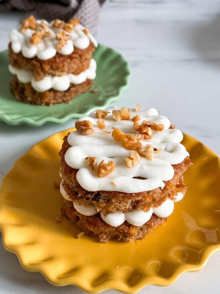 A beautifully plated mini carrot cake, sitting on a bright yellow plate with piped frosting and crunchy walnuts, while other cakes are arranged in the background.