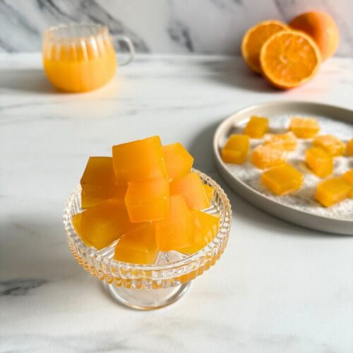 vegan fruit jellies on a cake stand, more fruit jellies on a plate in the back with sugar, oranges and a glass of orange juice, on a marble background