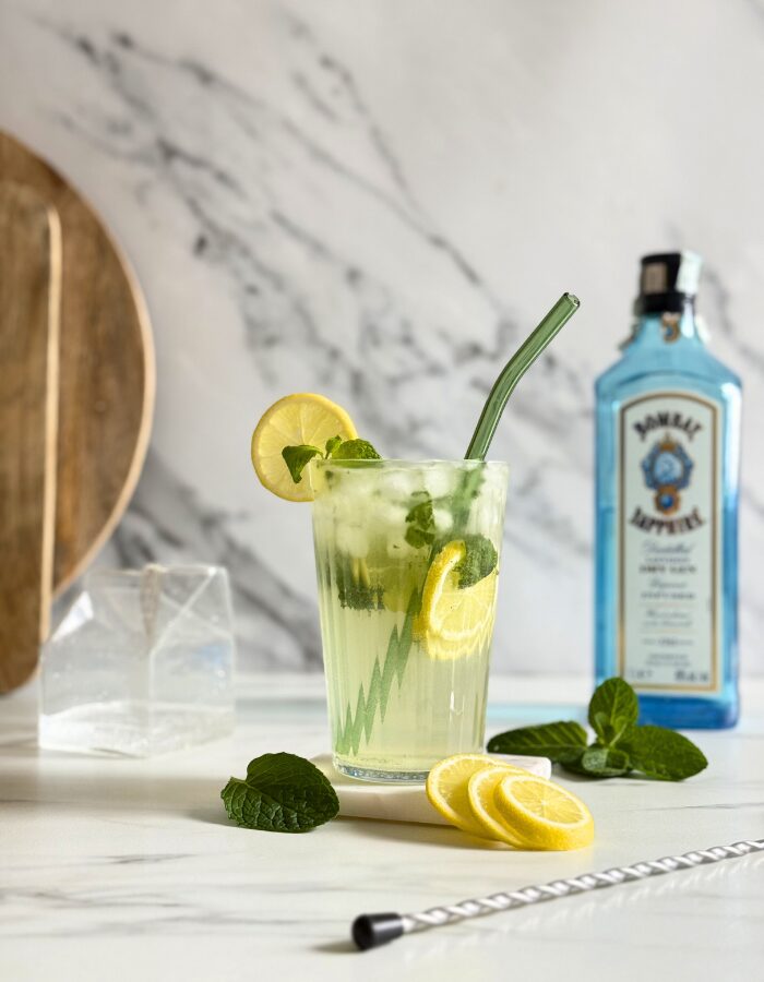 Easy Gin Mojitos - 5 Minute Twist On A Classic Mojito 7 gin mojito in a glass with a straw and mint and lemon, on a marble coaster, behind wooden boards, a bottle of sapphire gin and a container with club soda, on marble background