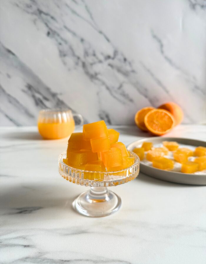 vegan fruit jellies on a cake stand, more fruit jellies on a plate in the back with sugar, oranges and a glass of orange juice, on a marble background