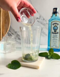 Easy Gin Mojitos - 5 Minute Twist On A Classic Mojito 12 ingredients being poured in the glass, on a marble coaster, behind wooden boards, a bottle of sapphire gin and a container with club soda, on marble background