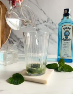 Easy Gin Mojitos - 5 Minute Twist On A Classic Mojito 13 ingredients being poured in the glass, on a marble coaster, behind wooden boards, a bottle of sapphire gin and a container with club soda, on marble background