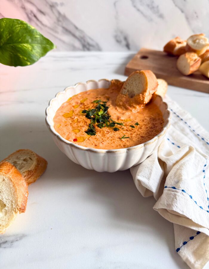 paprika dip in a bowl with a slice of bread, more bread on a wooden board behind and some on the side, a towel, a plant, on a marble background