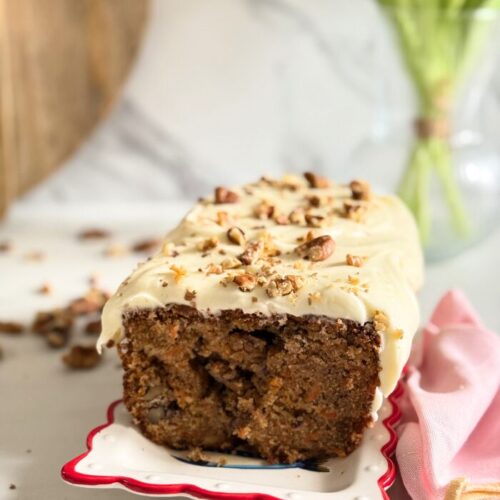 Delicious Carrot Cake Pound Cake With Cream Cheese Frosting 8 carrot cake pound cake on a plate, pin towel on the side, in the back a vase with tulips and wooden boards, walnuts around, on marble background