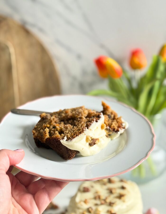 Delicious Carrot Cake Pound Cake With Cream Cheese Frosting 7 front space, a hand holding a plate with a slice of carrot cake, in the back carrot cake pound cake on a plate, pin towel on the side, in the back a vase with tulips and wooden boards, walnuts around, on marble background
