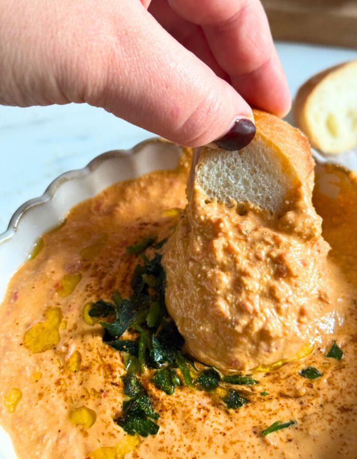 paprika dip in a bowl, fingers holding a slice of bread dipping in the dip