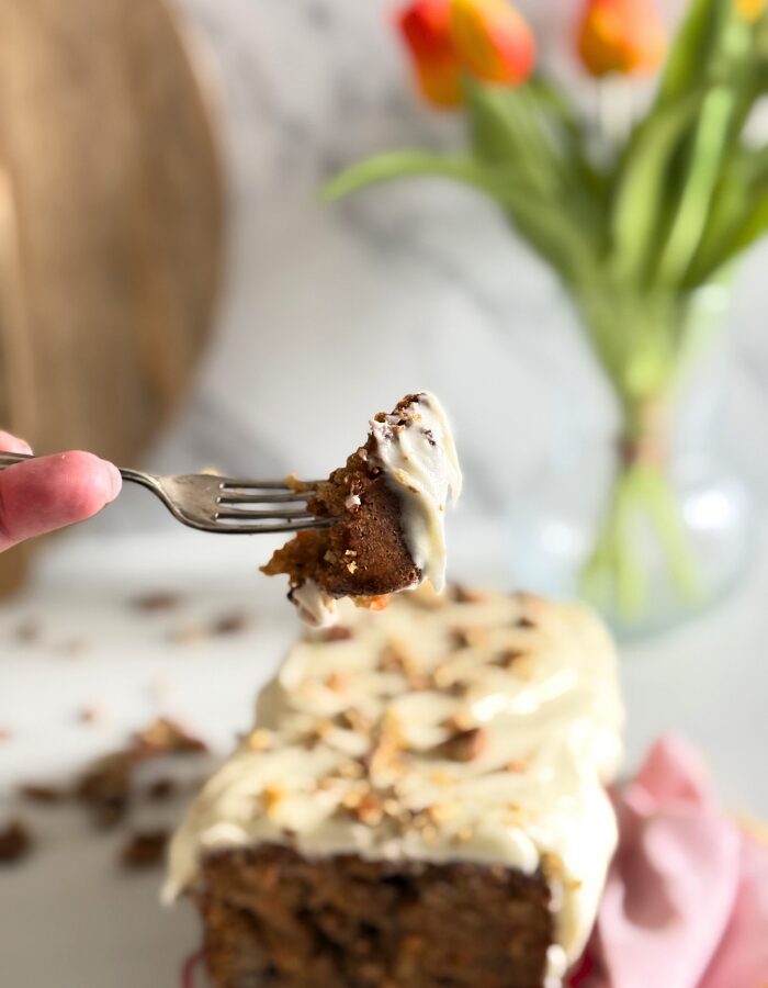 Delicious Carrot Cake Pound Cake With Cream Cheese Frosting 4 hand holding a fork with a bit of carrot cake pound cake, towel on the side, in the back a vase with tulips and wooden boards, walnuts around, on marble background
