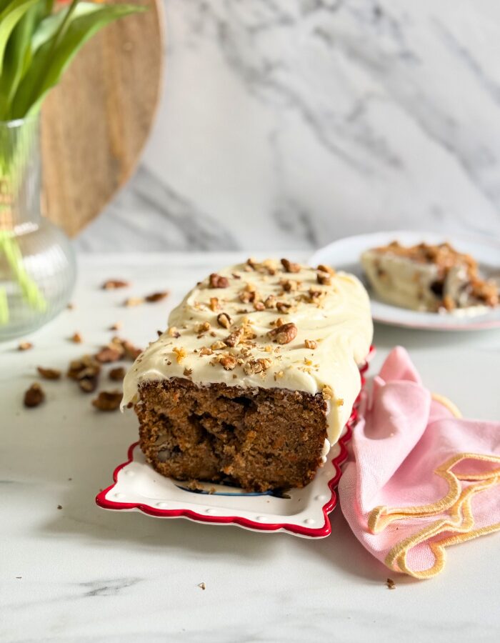Delicious Carrot Cake Pound Cake With Cream Cheese Frosting 3 carrot cake pound cake on a plate, pin towel on the side, in the back a vase with tulips, slice of cake on a plate, and wooden boards, walnuts around, on marble background