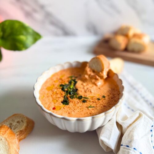 feta paprika dip in a bowl with a slice of bread, more bread on a wooden board behind and some on the side, a towel, a plant, on a marble background