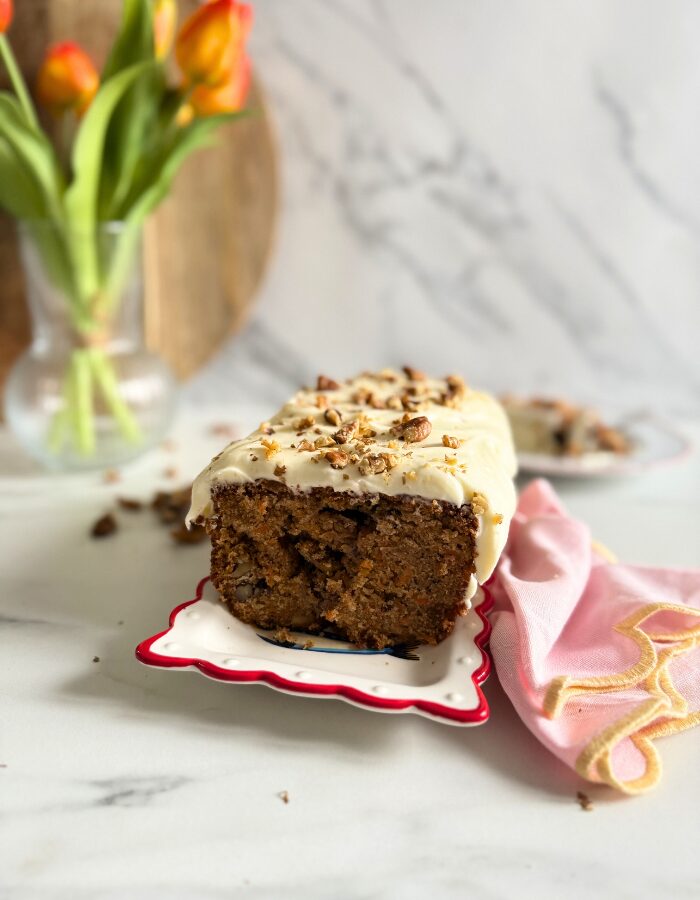 Delicious Carrot Cake Pound Cake With Cream Cheese Frosting 6 carrot cake pound cake on a plate, pin towel on the side, in the back a vase with tulips, slice of cake, and wooden boards, walnuts around, on marble background