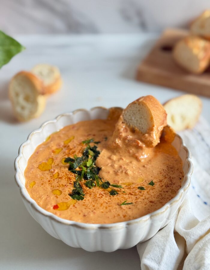 paprika dip in a bowl with a slice of bread, more bread on a wooden board behind and some on the side, a towel, a plant, on a marble background
