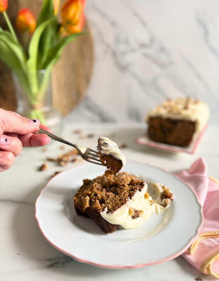 Delicious Carrot Cake Pound Cake With Cream Cheese Frosting 5 hand holding a fork with a bit of carrot cake pound cake, towel on the side, in the back a vase with tulips and wooden boards, walnuts around, on marble background