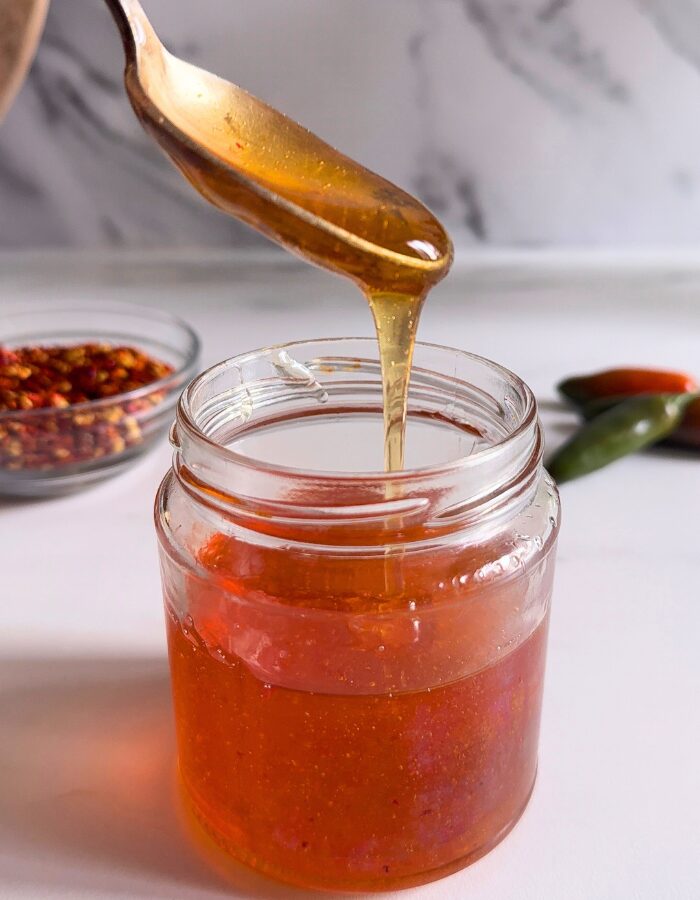 Easy Hot Honey Recipe - Quick Homemade Sauce 1 hot honey recipe in a jar, spoon holding some of it, behind a bowl with chilli flakes and green chilli peppers, on a marble background
