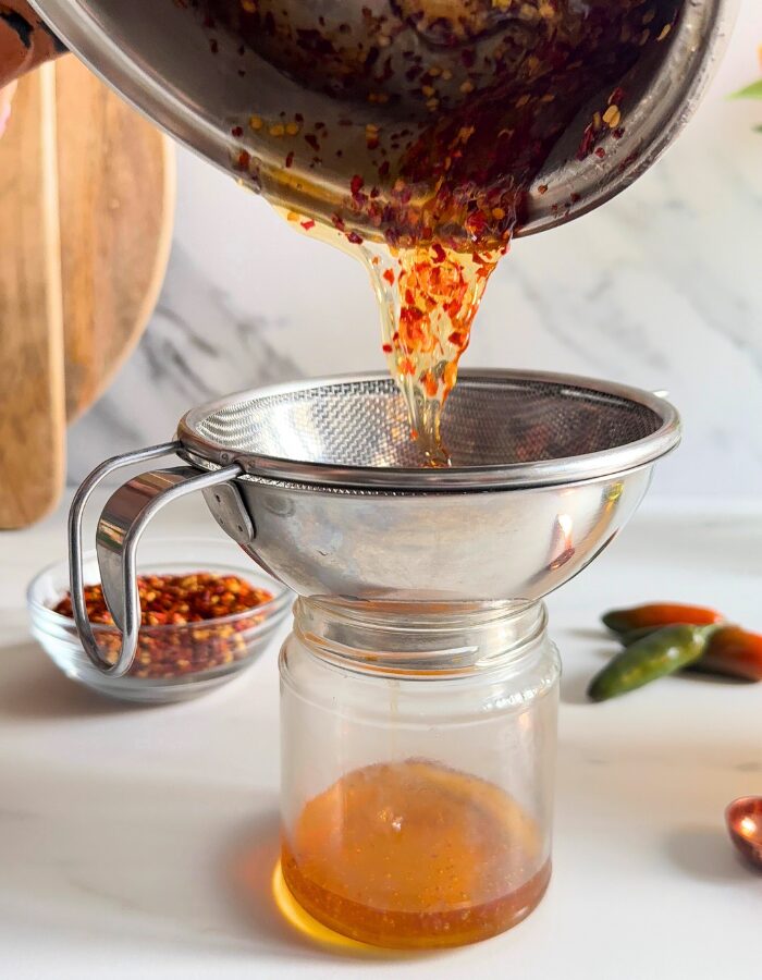 Easy Hot Honey Recipe - Quick Homemade Sauce 2 hot honey recipe being poured and strained in a glass container from a saucepan, chilli flakes in a bowl and chillies in the back, on marble background