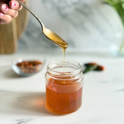 Easy Hot Honey Recipe - Quick Homemade Sauce 7 hot honey recipe in a jar, hand holding a spoon with some of it, behind a bowl with chilli flakes and green chilli peppers, on a marble background