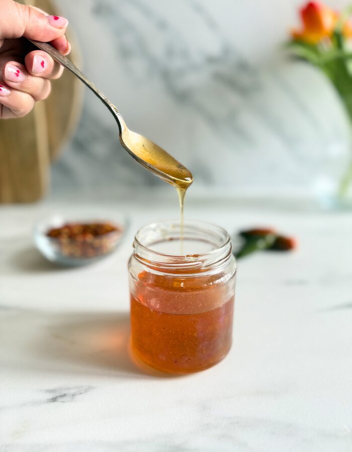 hot honey recipe in a jar, hand holding a spoon with some of it, behind a bowl with chilli flakes and green chilli peppers, on a marble background