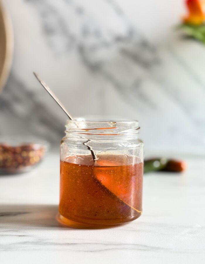 Easy Hot Honey Recipe - Quick Homemade Sauce 5 hot honey recipe in a jar, spoon holding some of it, behind a bowl with chilli flakes and green chilli peppers, on a marble background
