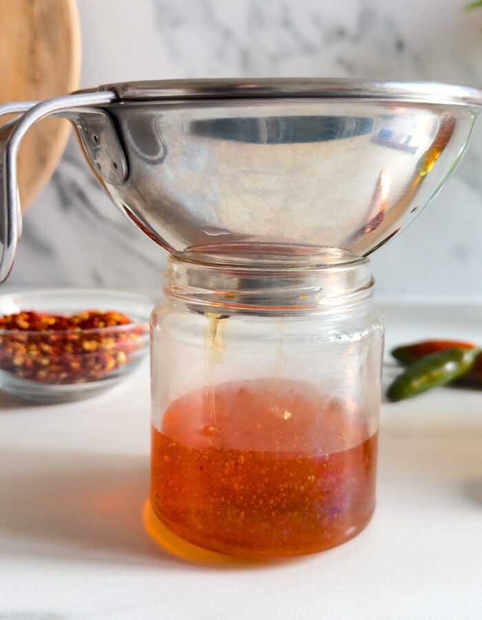Easy Hot Honey Recipe - Quick Homemade Sauce 3 hot honey recipe being poured and strained in a glass container, chilli flakes in a bowl and chillies in the back, on marble background
