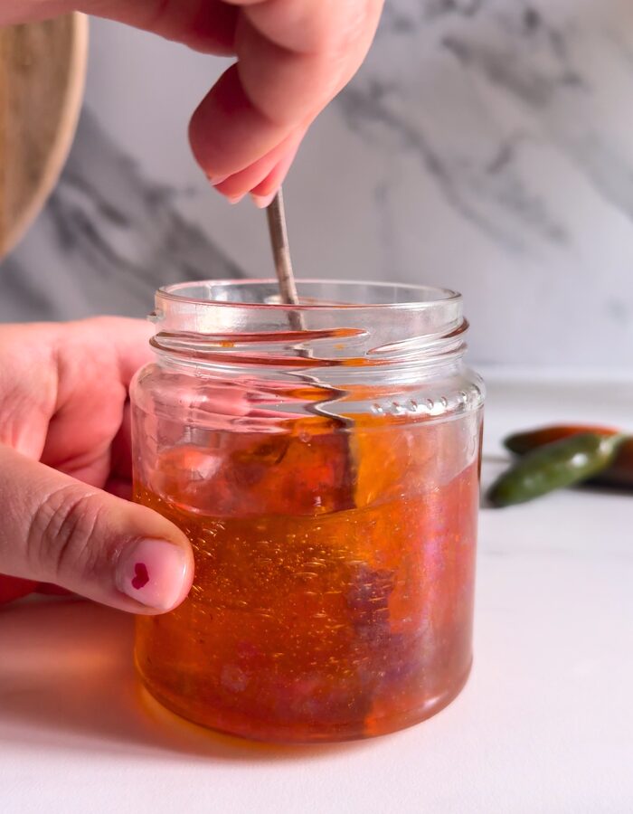 Easy Hot Honey Recipe - Quick Homemade Sauce 4 hands holding the jar and stirring hot honey recipe in a jar, behind a bowl with chilli flakes and green chilli peppers, on a marble background