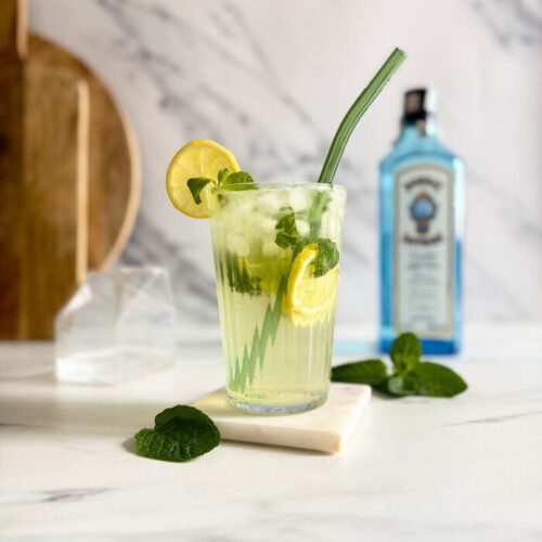 Easy Gin Mojitos - 5 Minute Twist On A Classic Mojito 10 gin mojito in a glass with a straw and mint and lemon, on a marble coaster, behind wooden boards, a bottle of sapphire gin and a container with club soda, on marble background