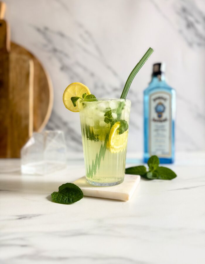Easy Gin Mojitos - 5 Minute Twist On A Classic Mojito 6 gin mojito in a glass with a straw and mint and lemon, on a marble coaster, behind wooden boards, a bottle of sapphire gin and a container with club soda, on marble background