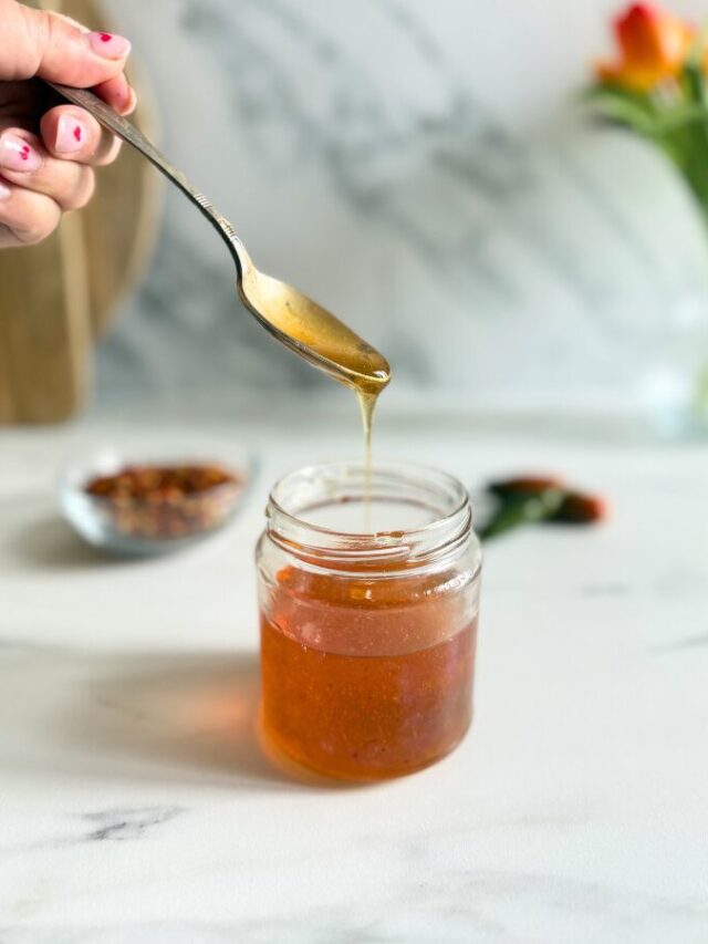 hot honey recipe in a jar, hand holding a spoon with some of it, behind a bowl with chilli flakes and green chilli peppers, on a marble background