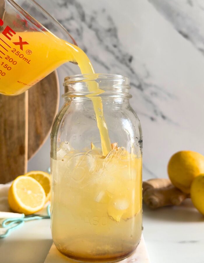 hand holding a jug of orange juice being poured into natures gatorade on a marble coaster, straws on front, lemons and ginger in the back with a bowl of salt