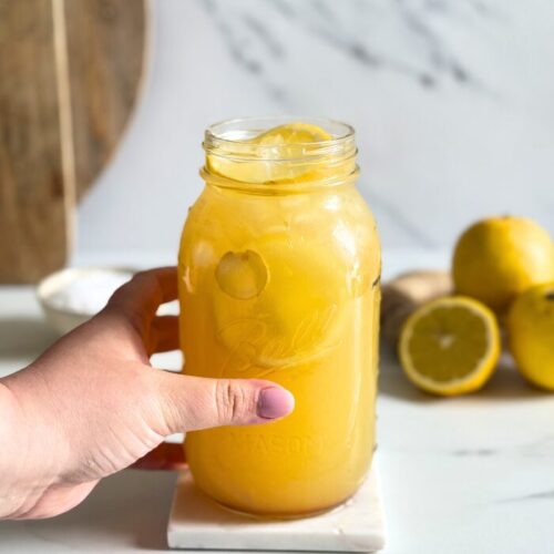 hand holding a jug of natures gatorade on a marble coaster, straws on front, lemons and ginger in the back with a bowl of salt