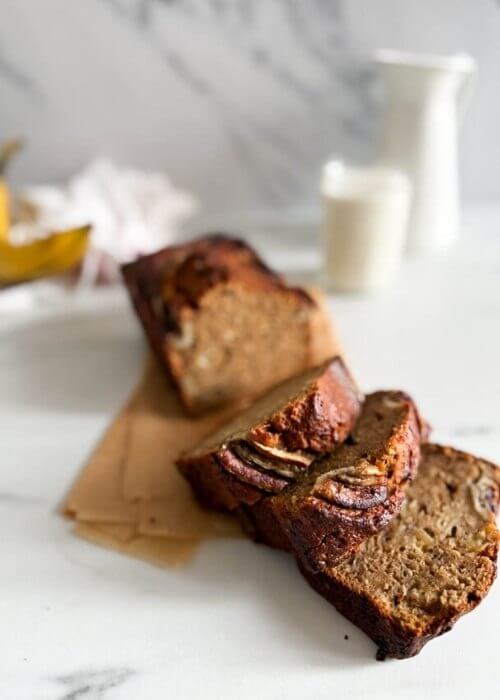 sliced 4 ingredient banana bread in a loafpan, on marble background, a glass of milk and towel on the side