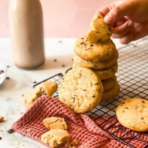 stack of SUGAR COOKIE WITH CHOCOLATE CHIPS on a wiring rack and pink towel, a hand holding one cookie, around a bottle of chocolate milk and chocolate chips